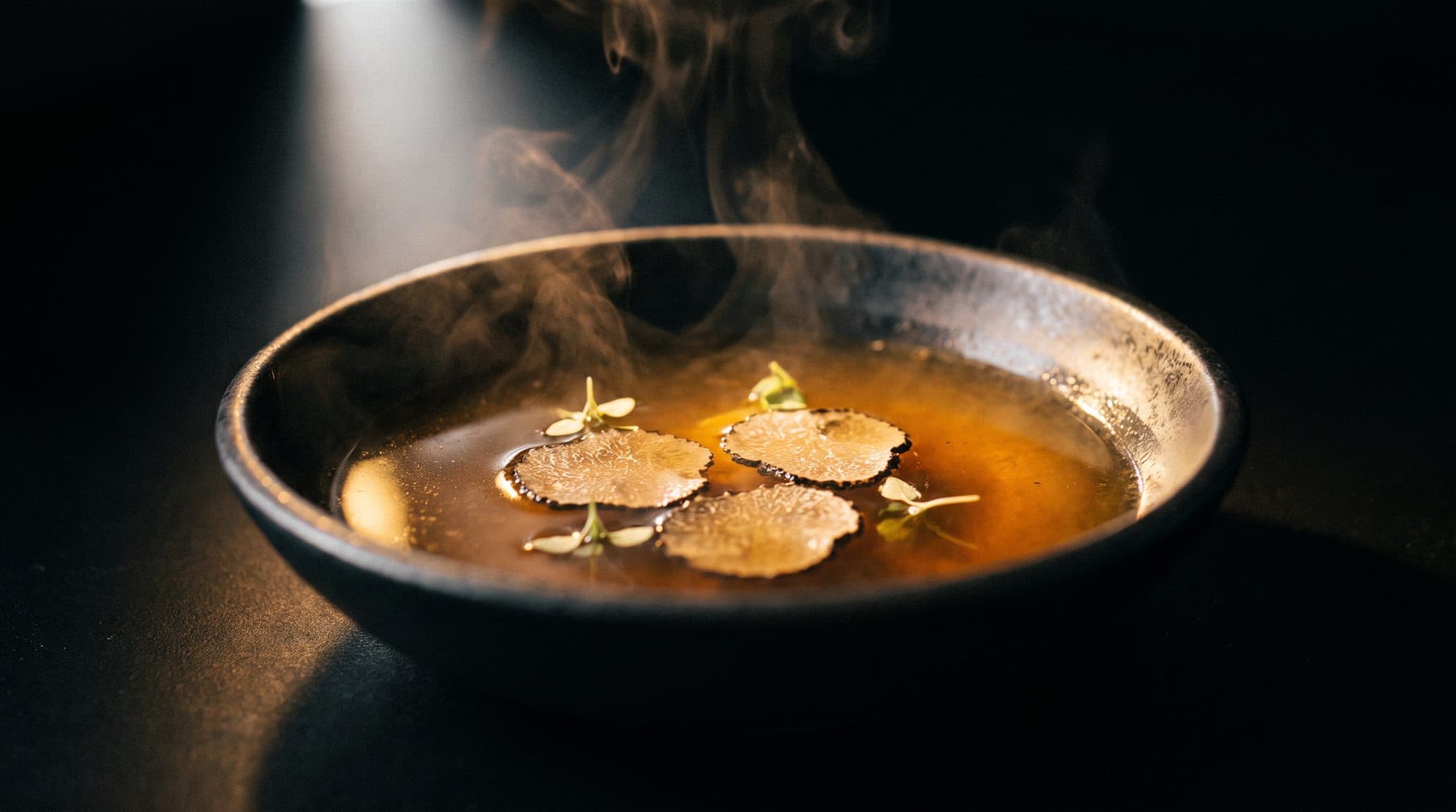 Un consommé ambré et limpide dans un bol sombre, quelques lamelles de truffe en surface, la vapeur qui monte.