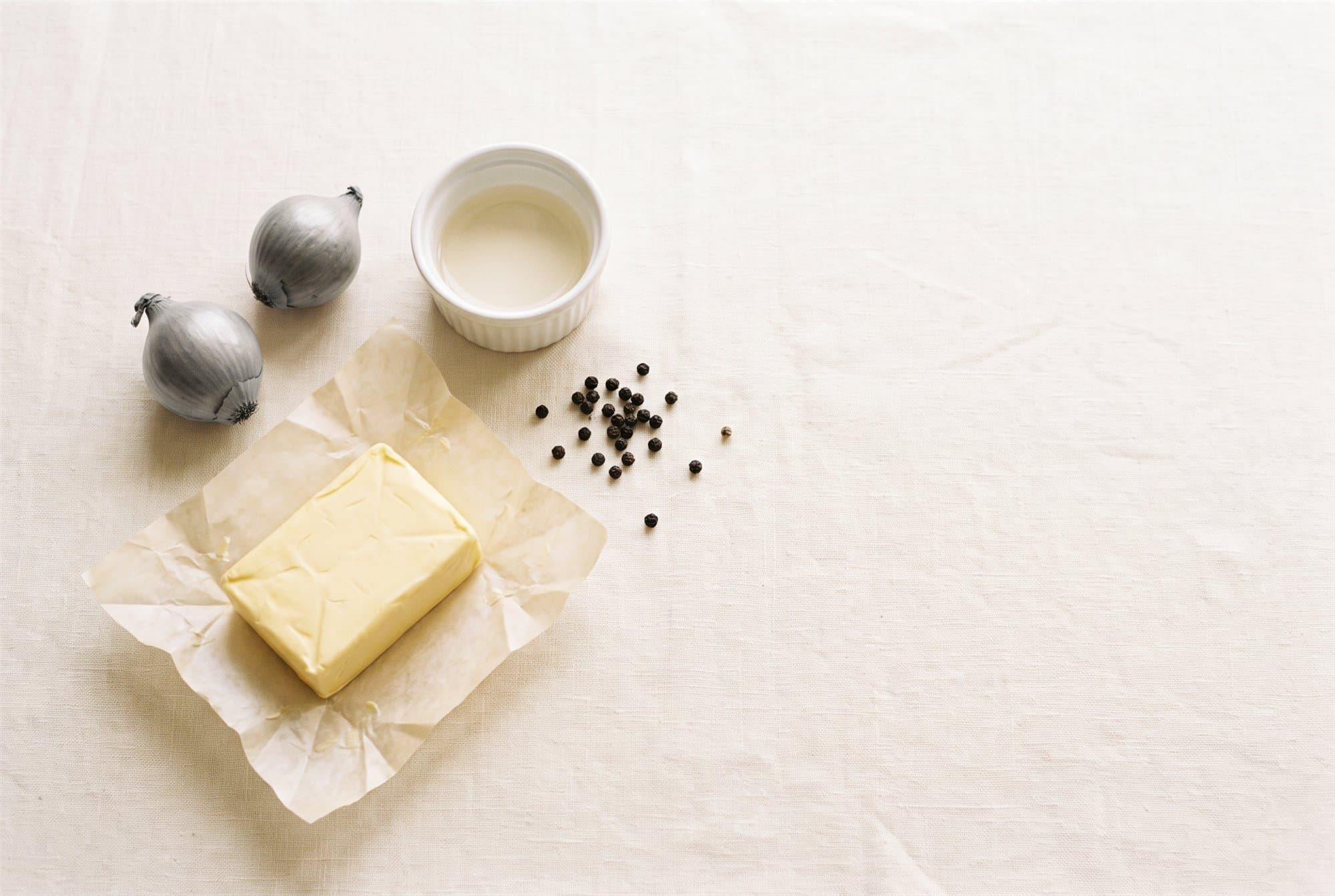 Deux échalotes grises, un beurre sur son papier, du poivre en grains et un ramequin de vinaigre blanc, posés sur une nappe de lin.